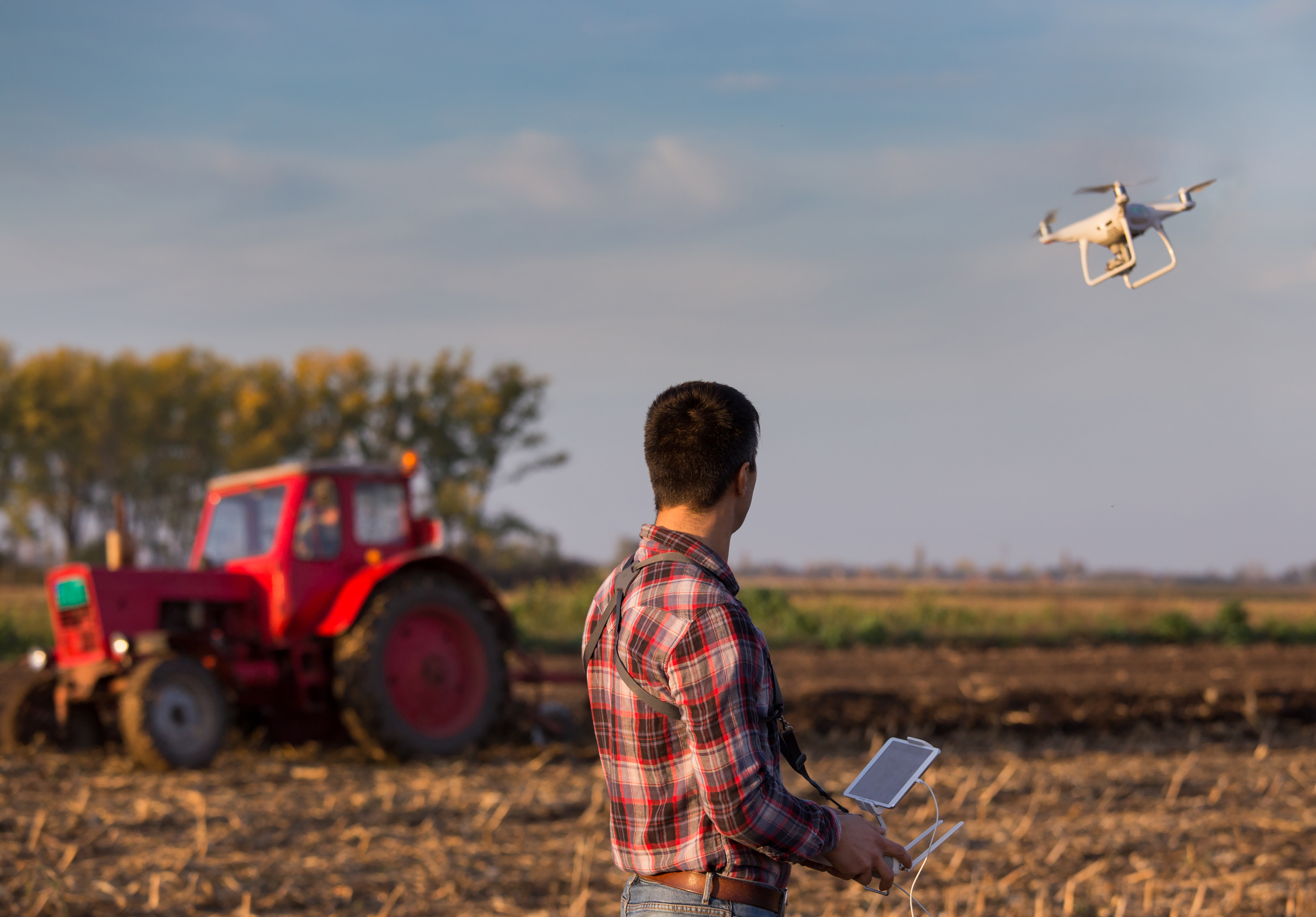 A person operates a drone over a tractor in a field 