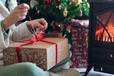 A person unties the ribbon on a gift box in front of a Christmas tree and a wood stove.