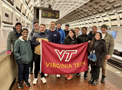 Students and faculty in Arlington pose for a picture in front of bookshelves while holding a Virginia Tech flag