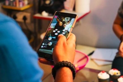 A hand holds a smartphone displaying a live video stream of someone decorating cupcakes. 
