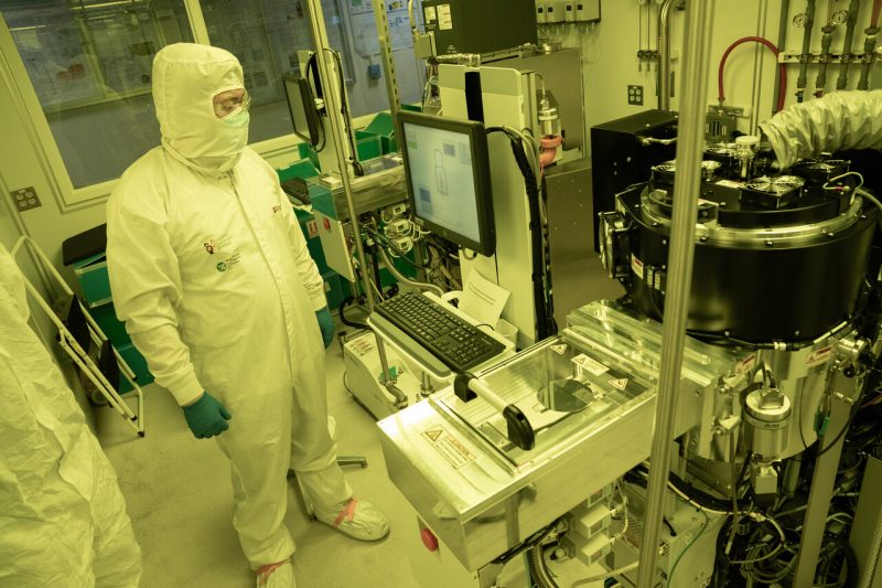 A person in a hood, mask, gloves, and protective boot covers standing in a yellow‑lit semiconductor lab while operating a machine with a monitor, keyboard, and connected equipment.