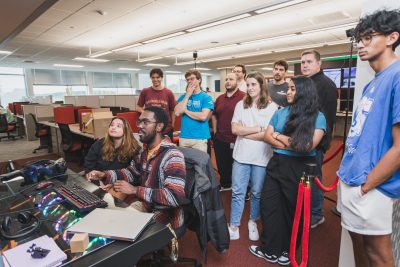 group of undergraduate students looking at screen, standing behind students at desk.